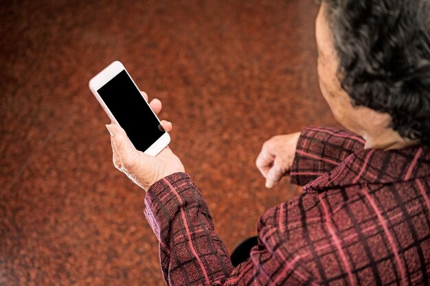 Mulher idosa asiática sentada e olhando através de algo no smartphone moderno fazendo conexão com outras pessoas em casa tecnologia de vida de perto