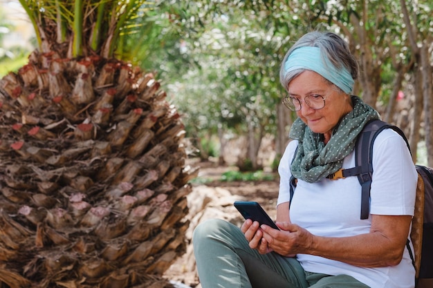 Mulher idosa ativa com mochila em caminhada ao ar livre, sentado à sombra de uma palmeira olhando para o celular mulher moderna idosa desfrutando de tecnologia e mídia social