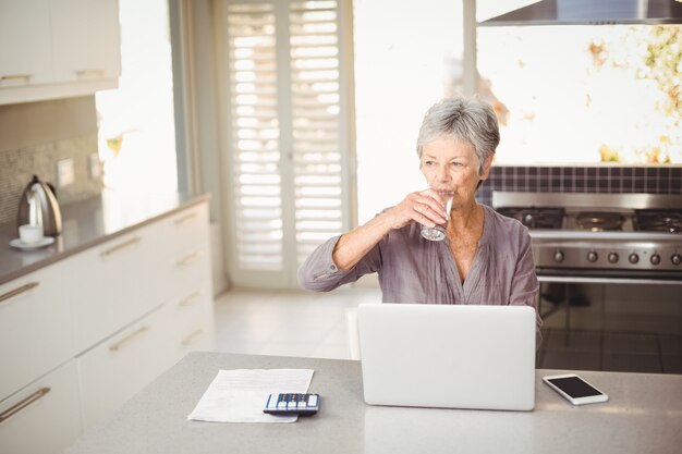 Mulher idosa bebendo água enquanto está sentado à mesa na cozinha