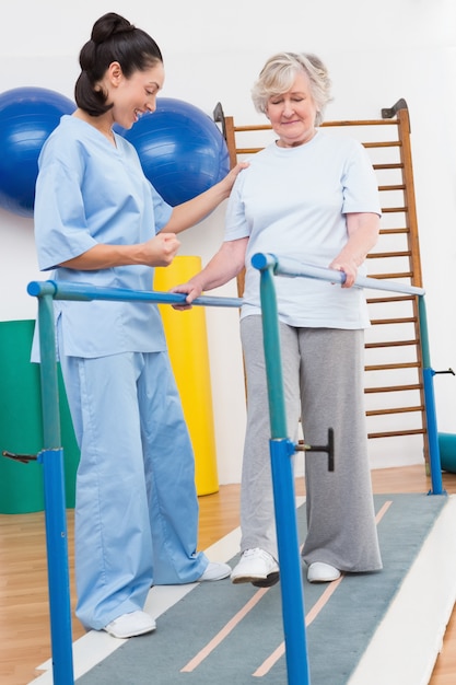 Foto mulher idosa caminhando com barras paralelas com terapeuta
