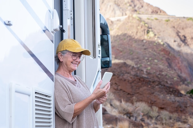 Mulher idosa caucasiana com chapéu amarelo do lado de fora de uma autocaravana segurando telefone celular senhora idosa desfrutando de estilo de vida alternativo