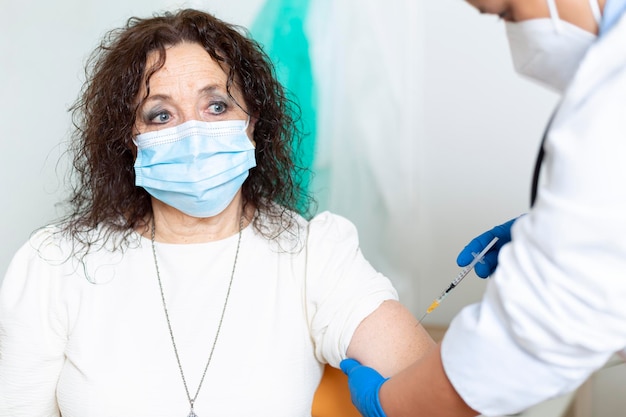 Mulher idosa, caucasiana, com máscara facial, sendo vacinada por um profissional de saúde. Campanha de vacinação contra o vírus Covid-19.