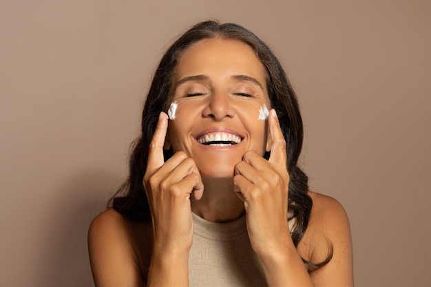 Foto mulher idosa caucasiana feliz aplica creme no rosto e desfruta de procedimentos de rotina e spa