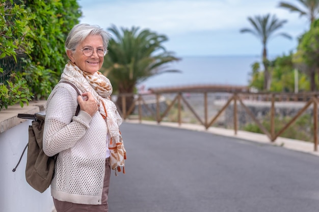 Mulher idosa caucasiana sorridente caminhando ao ar livre entre palmeiras e mar Mulher despreocupada sênior segurando mochila e olhando para a câmera