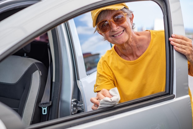 Mulher idosa caucasiana sorridente em camiseta amarela e chapéu limpa o carro em uma estação de lavagem de carros self-service cuidando dos detalhes olhando para a câmera Lavagem de carros self-service