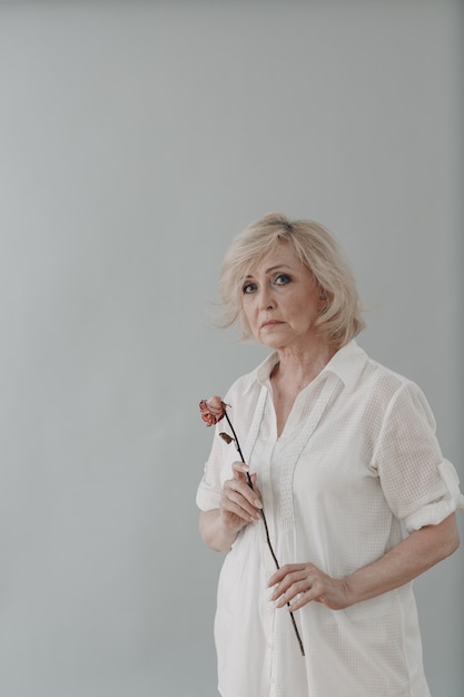 Mulher idosa chateada em vestido de branco segura solteira murcha e flor de rosa velha e seca