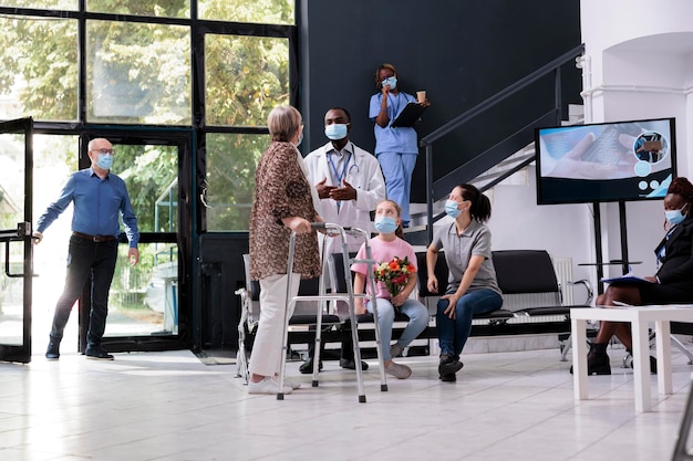 Mulher idosa com andador discutindo diagnóstico médico com médico durante visita de check-up na área de espera do hospital. Pessoas usando máscara facial protetora para prevenir infecção por coronavírus