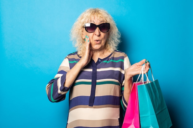 Mulher idosa com cara de surpresa segurando sacolas de compras na superfície azul