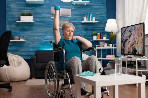 Mulher idosa com deficiência em cadeira de rodas levantando braço, treinamento de resistência de músculos usando recuperação de halteres após deficiência muscular assistindo a um vídeo cardiovascular no laptop