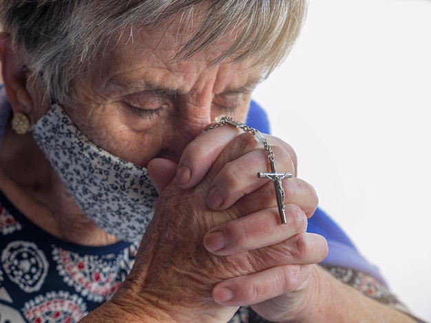 Mulher idosa com máscara protetora contra o vírus orando com os olhos fechados e um crucifixo nas mãos entrelaçadas. conceito de cristianismo