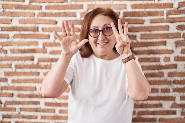 Foto mulher idosa com óculos de pé sobre a parede de tijolos mostrando e apontando para cima com os dedos número oito enquanto sorri confiante e feliz