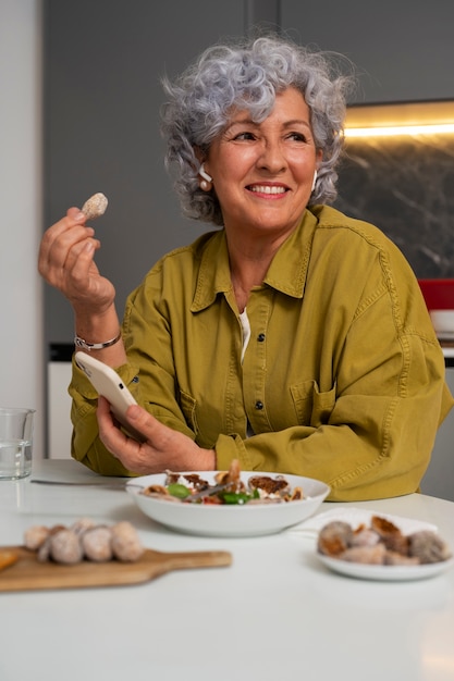 Mulher idosa comendo prato de figos em casa e usando smartphone