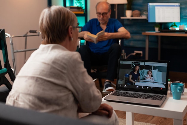 Mulher idosa conversando com parentes em videoconferência com o laptop em casa. sobrinha doente ligando para a avó do hospital com a mãe na internet online. homem em cadeira de rodas no fundo