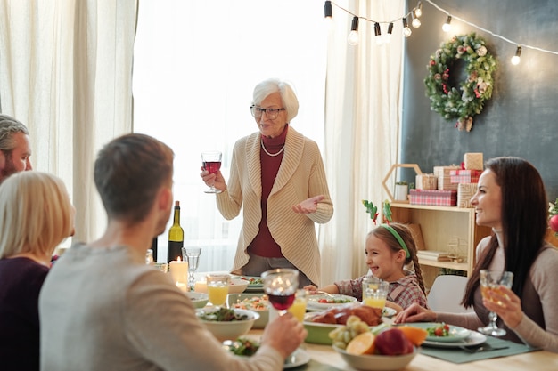 Mulher idosa de cabelos grisalhos brindando com uma taça de vinho tinto servida na mesa em frente à família durante o jantar