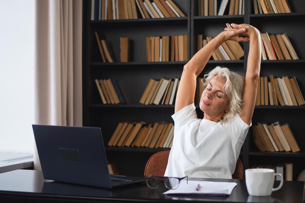 Mulher idosa de meia-idade relaxando, esticando as mãos e o corpo, fazendo uma pausa do trabalho no laptop em casa