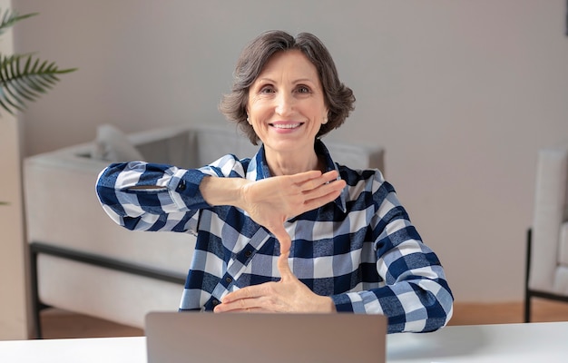 Mulher idosa e surda feliz usa linguagem de sinais durante uma videochamada usando um laptop enquanto está sentada no escritório em casa