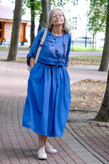 Mulher idosa elegante de vestido azul e óculos caminha pelas vielas do parque