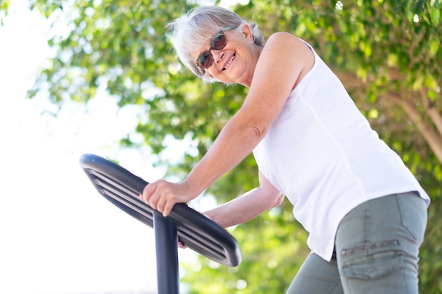 Mulher idosa em atividade esportiva em parque público, sentada no equipamento de bicicleta, fazendo exercícios, sorrindo