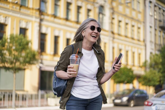 Mulher idosa europeia com cabelos grisalhos feliz aproveitando o tempo livre depois do trabalho ou viajando segurando o telefone enquanto toma suco em qualquer lugar usando um copo de plástico no fundo da cidade aproveitando a vida mulher madura