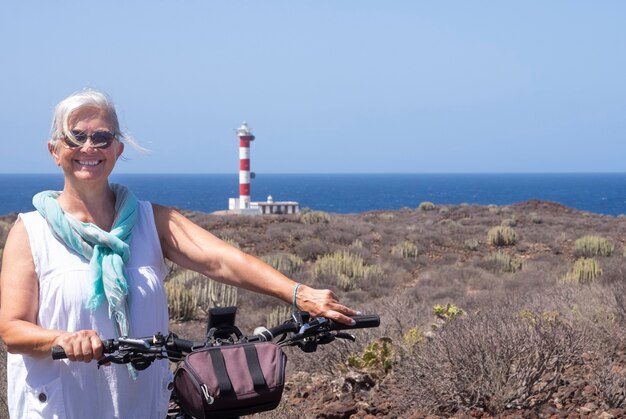 Mulher idosa feliz com cabelo branco e óculos, curtindo a liberdade e passeios de bicicleta perto de um farol - mar azul e horizonte sobre a água em um dia ensolarado em Tenerife, Ilhas Canárias