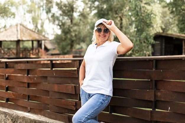 Mulher idosa feliz em um boné com óculos de sol em uma camiseta branca com jeans azul fica perto de uma cerca de madeira no jardim rural