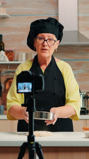 Mulher idosa feliz padeiro filmando vlog de culinária na cozinha de casa. influenciador chef de blogueiro aposentado que usa tecnologia da internet para se comunicar, fazer blogs nas redes sociais com equipamento digital