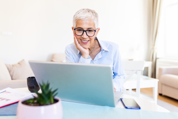 Mulher idosa focada com cabelos brancos em casa usando laptop Empreendedor sênior elegante usando óculos trabalhando em computador em casa Mulher analisando e gerenciando contas domésticas e finanças domésticas