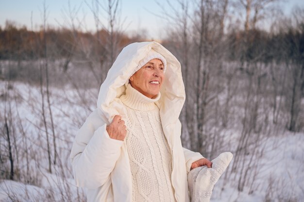 Mulher idosa idosa feliz com roupas brancas quentes brincando com a neve no inverno ensolarado ao ar livre