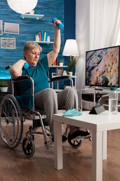 Mulher idosa inválida segurando halteres de treino levantando o braço enquanto exercita a persistência dos músculos do corpo fazendo exercícios cardiovasculares