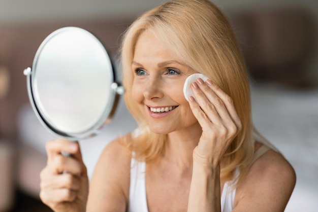 Mulher idosa limpando a pele com almofada de algodão olhando para o espelho e sorrindo para seu reflexo desfrutando