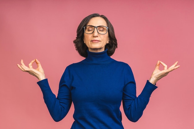 Mulher idosa madura sênior sorridente de mãos dadas em gesto de ioga relaxante meditando, tentando se acalmar isolado no retrato de estúdio de fundo de cor rosa.