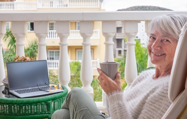 Mulher idosa madura sorridente relaxando na varanda de casa enquanto usa laptop para trabalho remoto mulheres adultas atraentes desfrutando de um estilo de vida pacífico ao ar livre bebendo um café