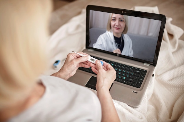 Mulher idosa na cama, olhando para a tela do laptop e consultando um médico online em casa, serviços de telessaúde durante o bloqueio, videochamada distante, aplicativo de saúde de tecnologia moderna