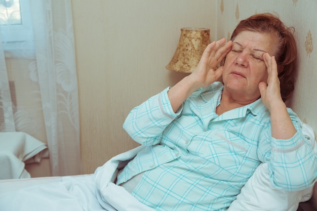 Mulher idosa se sentindo mal na cama, segurando as mãos na cabeça. Dor de cabeça pela manhã.