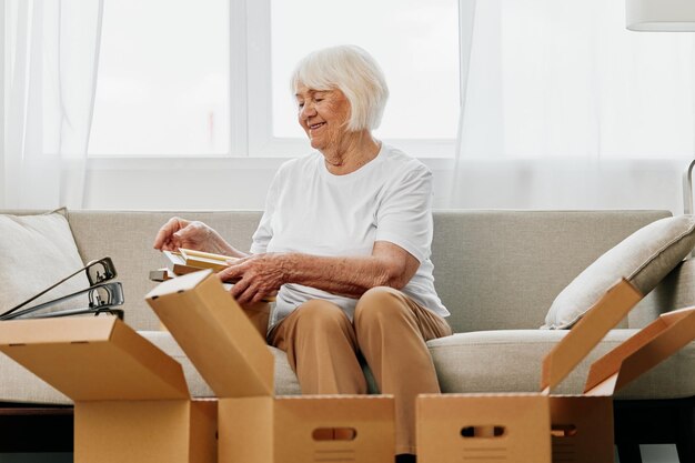 Mulher idosa senta-se em um sofá em casa com caixas coletando coisas com álbuns de memórias com fotos e molduras mudando para um novo lugar limpando as coisas e um sorriso feliz Aposentadoria do estilo de vida