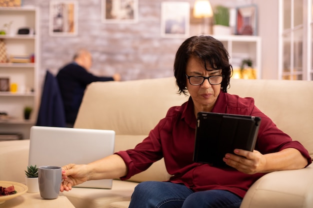 Mulher idosa sentada no sofá e usando um tablet Pc digital na aconchegante sala de estar.