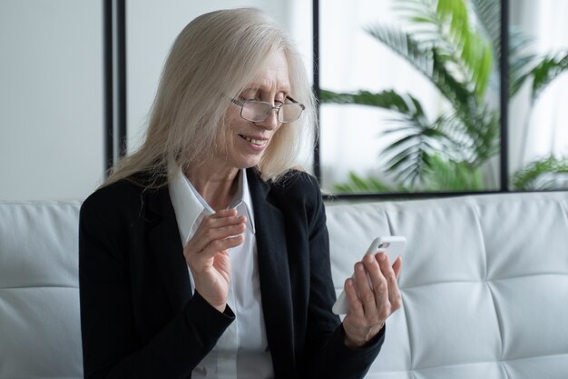 Mulher idosa sentada no sofá sorrindo enquanto usa o telefone comemorando o sucesso