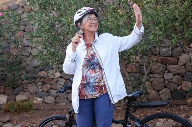 Mulher idosa sorridente com capacete em atividade ao ar livre com bicicleta elétrica agitando a mão