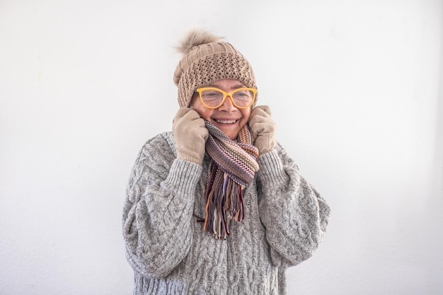 Mulher idosa sorridente de suéter cinza com óculos e boné cobre o pescoço porque sente frio isolado no fundo branco