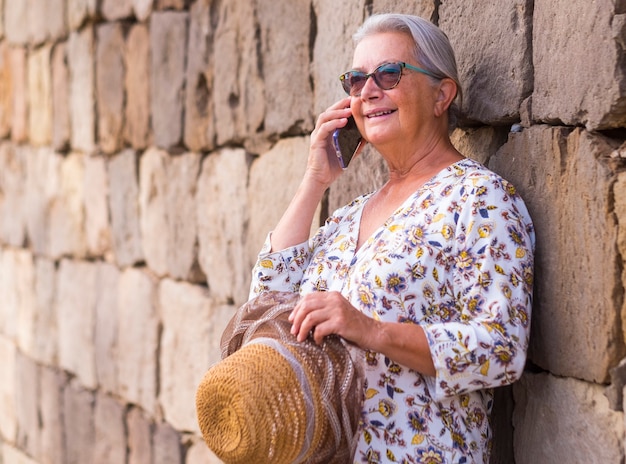 Mulher idosa sorridente e relaxada falando com um telefone inteligente em pé contra uma parede de pedra com óculos de sol