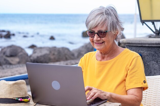 Mulher idosa sorridente, sentada ao ar livre na mesa do café usando o laptop. Trabalhador nômade blogueiro ativo
