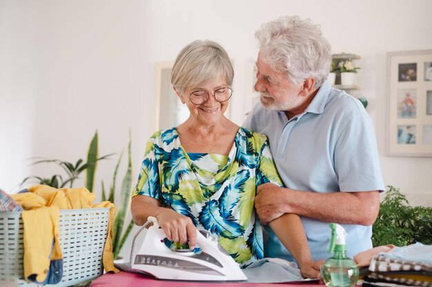 Mulher idosa sorrindo enquanto passa em casa na tábua de passar roupa enquanto seu marido olha para seu casal de idosos ternamente caucasianos na aposentadoria