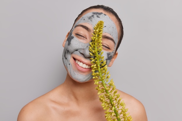 Mulher inclina a cabeça mantém os olhos fechados sorri amplamente mostra dentes brancos gosta de rotina de cuidados com a pele aplica máscara nutritiva de argila para remoção de poros de cravos pretos mantém poses de flores silvestres sem camisa dentro de casa