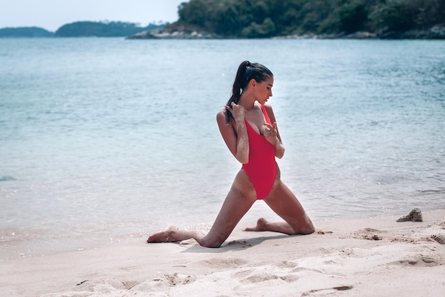 Mulher incrível, aproveitando o sol e as ondas em uma praia branca. a morena está ajoelhada na água em um maiô sexy vermelho vista de cima.