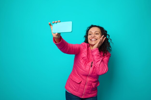 Mulher incrível de cabelos escuros atraente com penteado encaracolado, vestindo jaqueta rosa, segurando a mão perto do rosto, sorri sorriso cheio de dentes olhando para a câmera do smartphone, fazendo selfie. Espaço de cópia de fundo azul