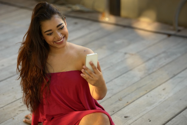 Mulher indiana bonita jovem feliz usando o telefone enquanto está sentado no chão de madeira