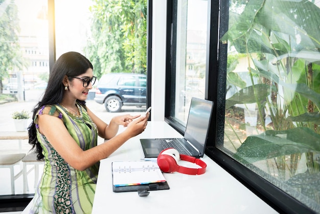 Mulher indiana com óculos usando um telefone celular com um laptop e um notebook em cima da mesa