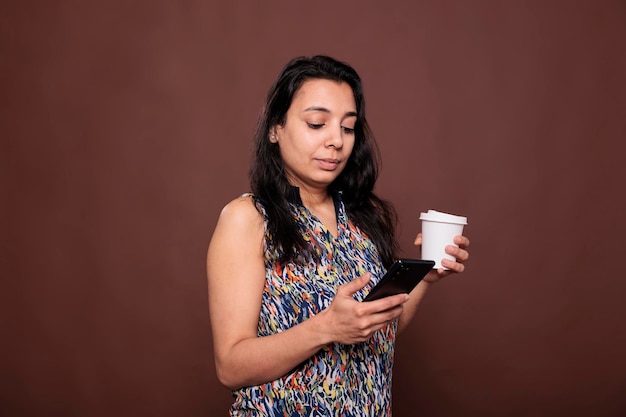 Mulher indiana conversando em redes sociais, tomando café, usando smartphone, enviando mensagens de texto. Pessoa segurando chá para ir copo de papel, mensagens de texto no telefone, plano médio de estúdio