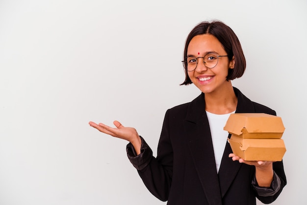 Mulher indiana de negócios jovem comendo hambúrgueres isolados, mostrando um espaço de cópia na palma da mão e segurando a outra mão na cintura.