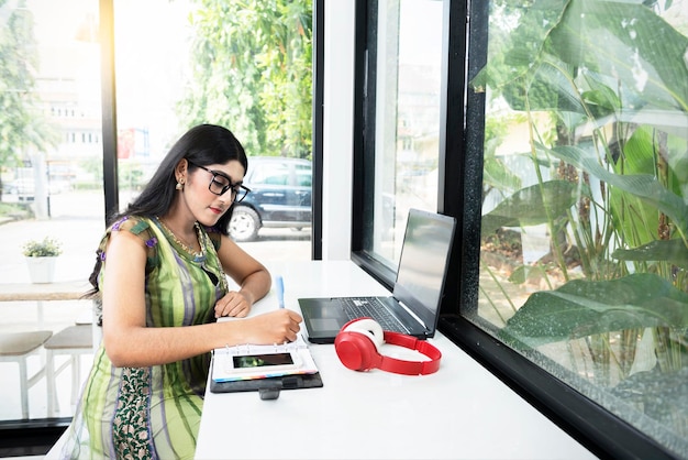 Mulher indiana em óculos escrevendo algo em um notebook com um laptop e celular em cima da mesa